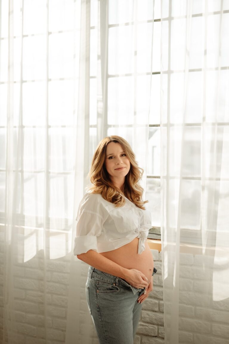 Těhotná žena pózuje vedle okna při těhotenském focení v ateliéru Praha